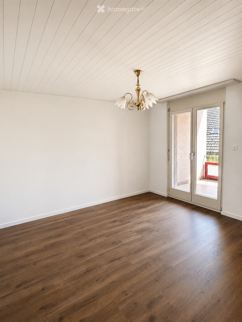 Empty room with wooden flooring, white walls, and a chandelier hanging from the ceiling. A large sliding glass door leads to an outdoor area.