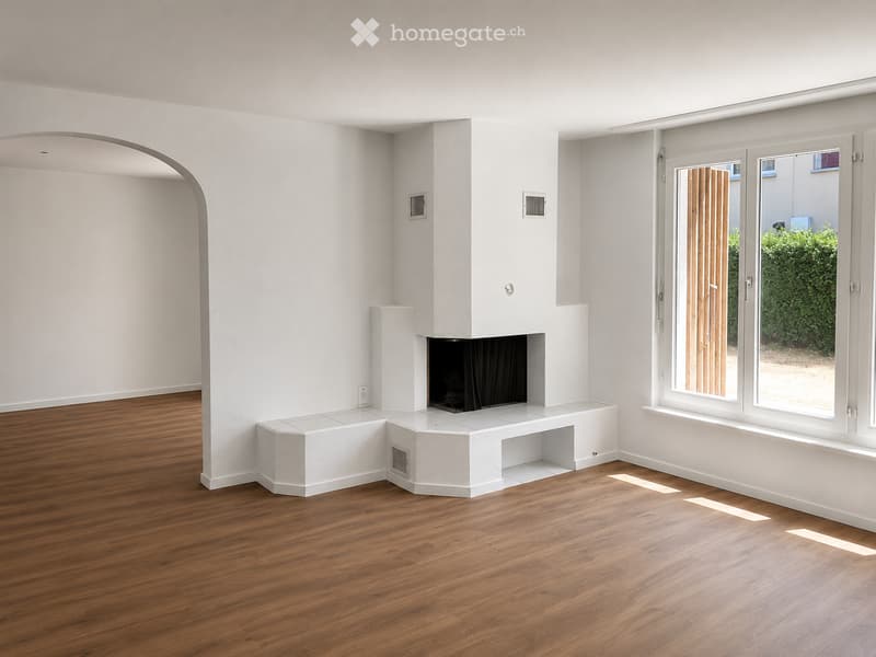 Empty room with wooden flooring, white walls, and a built-in fireplace. Large windows and sliding doors provide natural light and access to the outdoors.