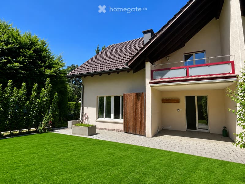 Two-story house with a red-tiled roof, white walls, large windows, and a balcony on the upper floor. The front yard features a well-maintained lawn, a brick pathway, and a small planter with greenery.