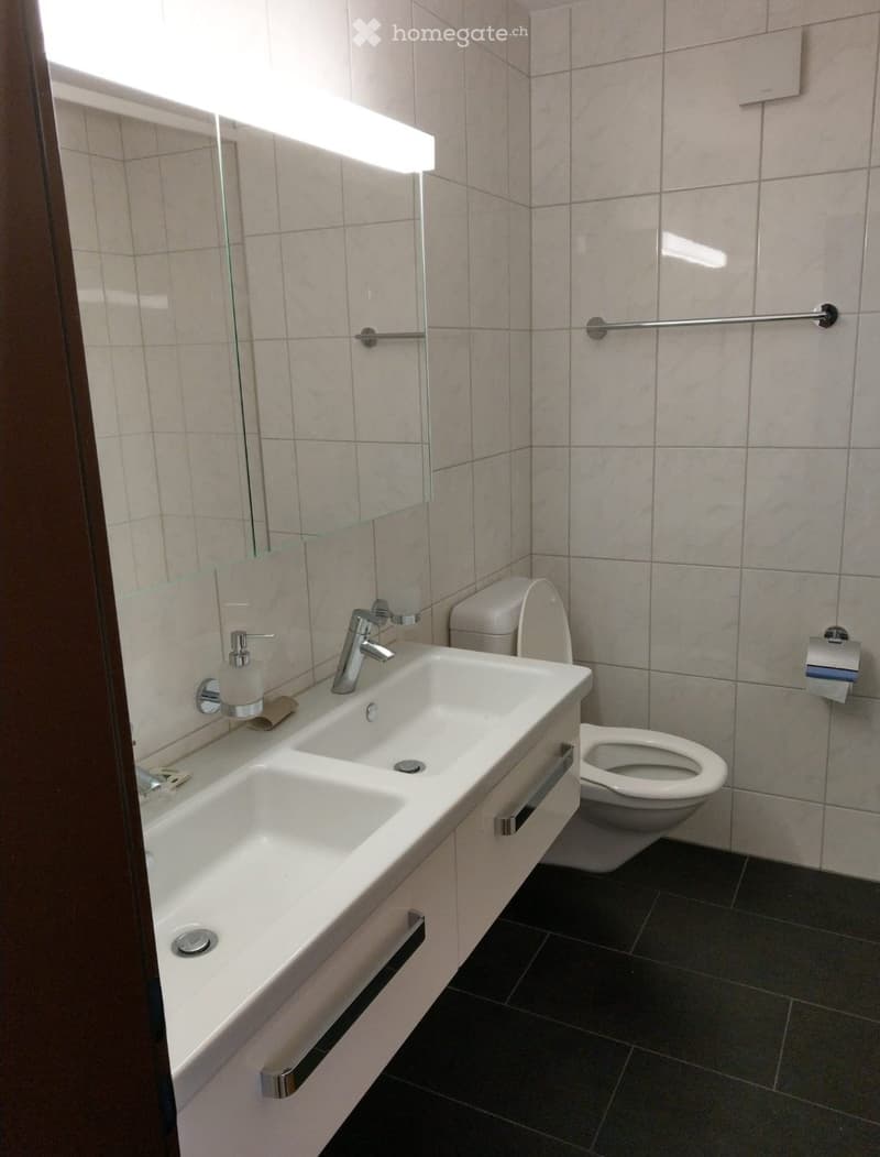 Modern bathroom with a white sink, chrome faucet, toilet, and a large mirror with built-in lighting. The walls are tiled in light beige, and the floor is dark grey.