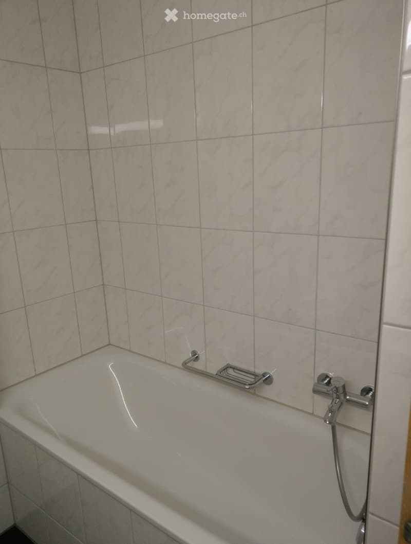 Bathtub with a modern chrome handrail and faucet, surrounded by light-colored tiled walls.