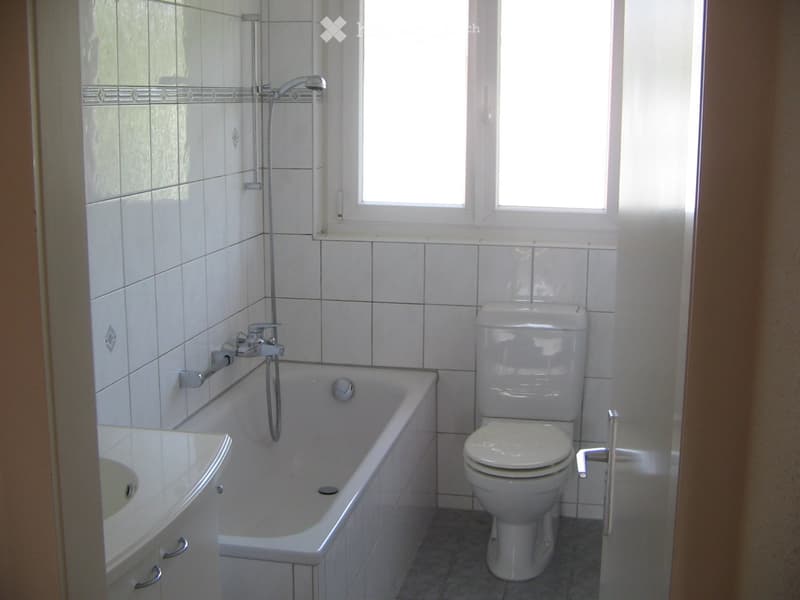 Bathroom with white tiles, bathtub, toilet, sink, and a window above the bathtub.
