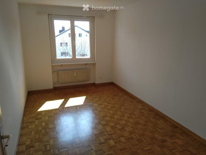 Empty room with wooden flooring, white walls, and a window with a view of neighboring buildings.