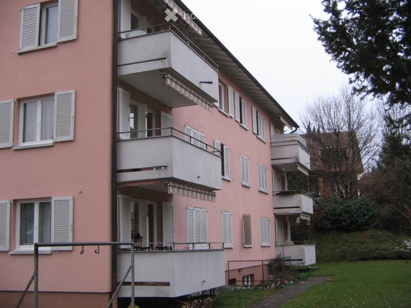 Pink multi-story apartment building with multiple balconies, white shutters, and a small garden area in front.