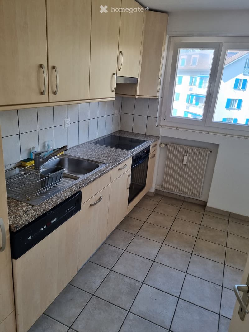 Modern kitchen with light wood cabinets, granite countertop, stainless steel sink, oven, and a window overlooking a building.