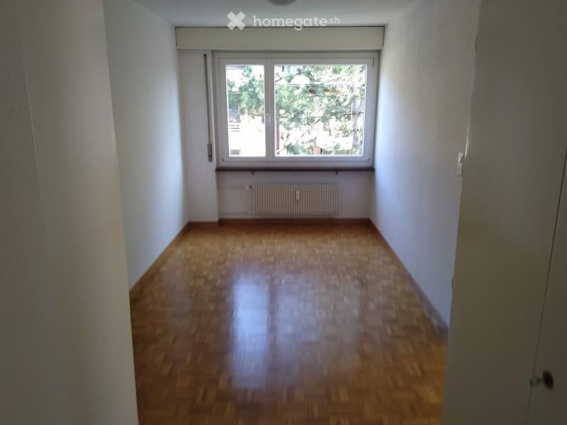 Empty room with wooden flooring, white walls, and a large window with a view of trees and a building outside. A radiator is visible below the window.