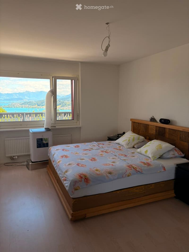 Cozy bedroom with wooden bed, floral bedding, nightstand, radiator, and air conditioner. Large window offers scenic mountain and lake view.
