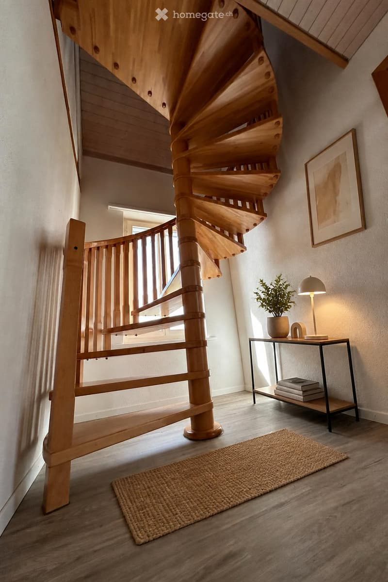 Spiral wooden staircase with a modern design, accompanied by a small side table with a lamp, a potted plant, and a woven rug on the floor.