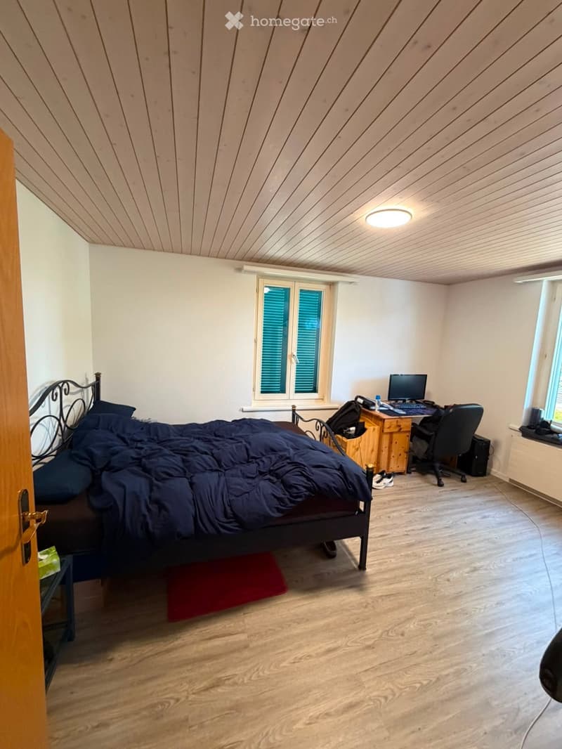 Cozy attic bedroom with wooden floors, slanted wooden ceiling, large windows with shutters, a bed with dark bedding, a desk with a computer, and a chair.