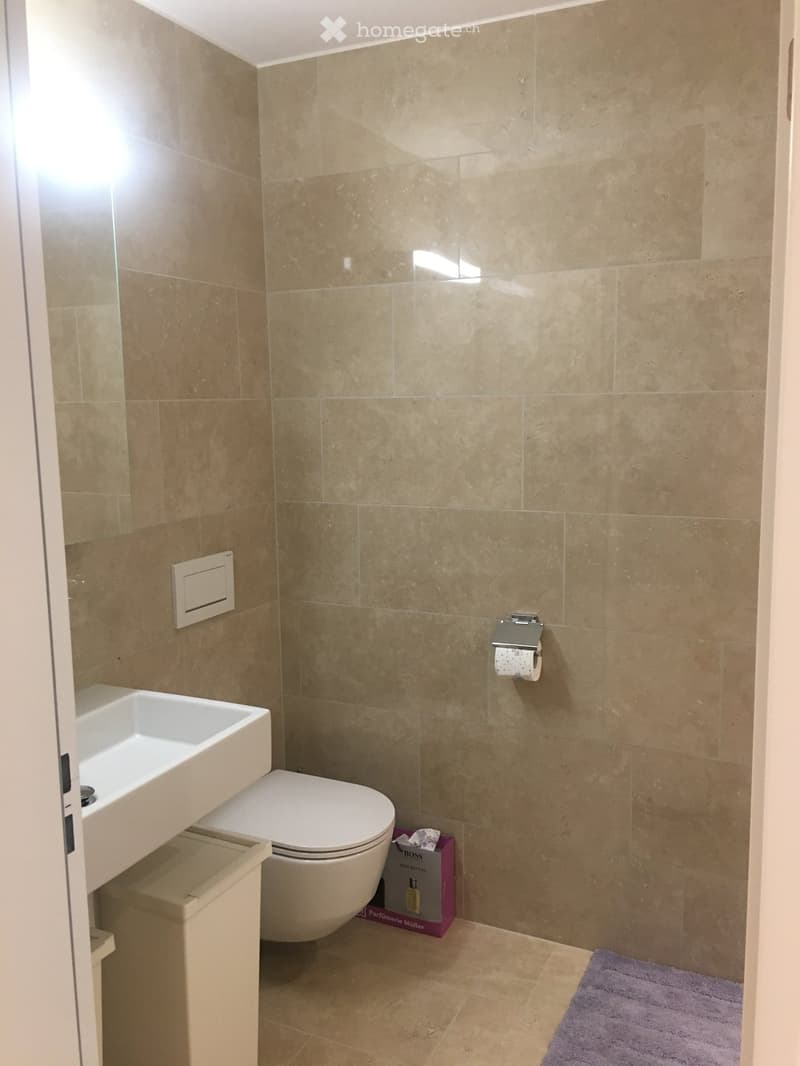 Modern bathroom with beige tiles, white toilet, sink, and a purple rug on the floor.