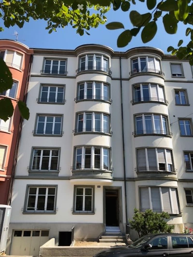 Multi-story residential building with white facade, multiple windows, balconies, and a garage entrance at the bottom.