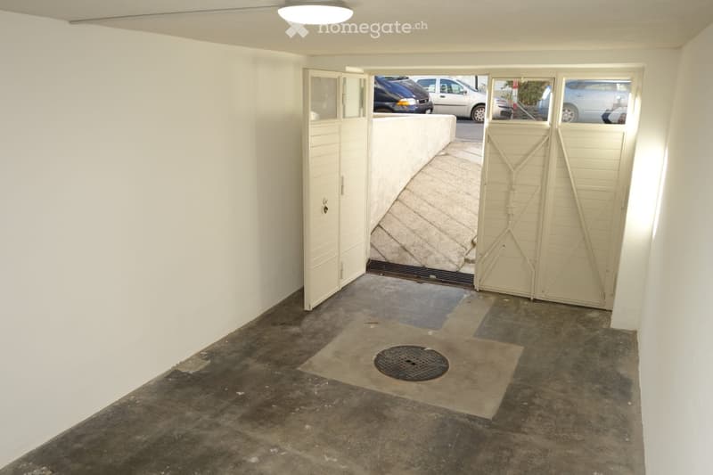 Empty room with white walls, concrete floor, and a large open doorway leading to an outdoor area with parked cars visible.