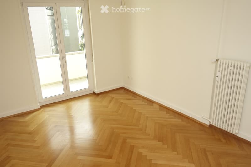Empty room with light-colored wooden herringbone flooring, white walls, and a white radiator. A large glass sliding door leads to a balcony or outdoor area.