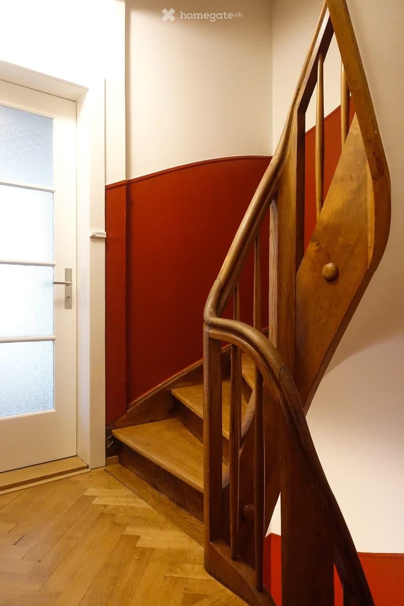 Spiral staircase with wooden steps and a metal railing, leading up from a wooden floor area. The walls are painted in a two-tone design with white upper sections and red lower sections.