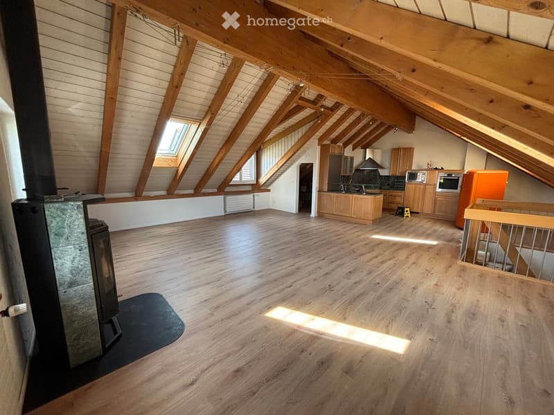 Open-plan attic space with exposed wooden beams, wooden flooring, and a modern kitchen area. Features a fireplace and skylights.