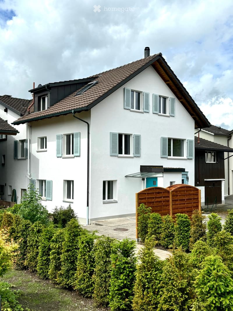 Two-story house with white exterior, light blue shutters, and a tiled roof. Features a wooden gate, small garden, and a blue front door.