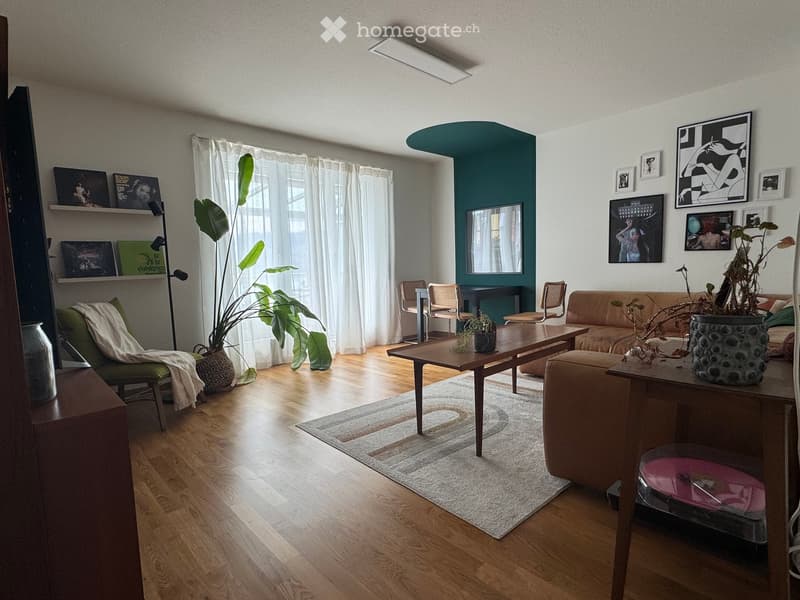 Modern living room with wooden flooring, a green accent wall, large windows with white curtains, a beige sofa, a wooden coffee table, a green armchair, and decorative shelves with artwork.