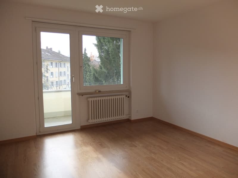 Empty room with wooden flooring, large window, sliding glass door leading to a balcony, radiator, and visible light fixture.