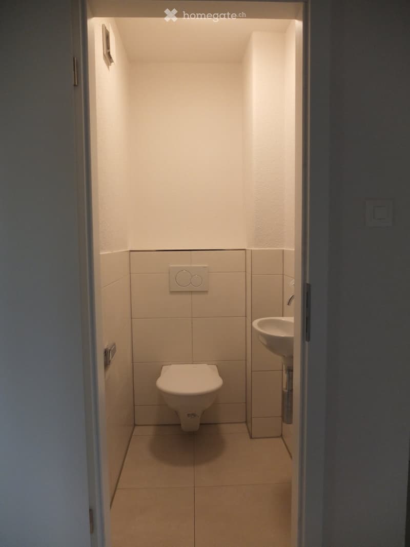 Modern, compact bathroom with white tiled walls and floor, featuring a toilet and a sink.