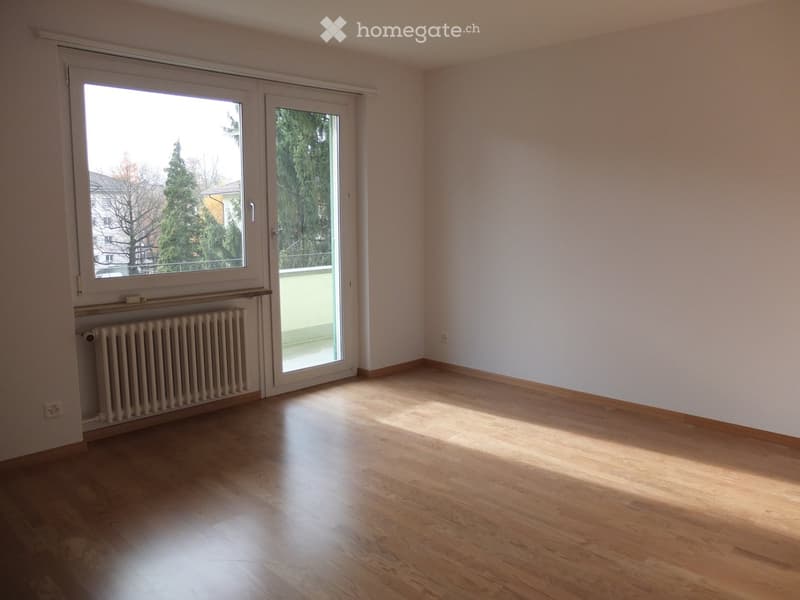 Empty room with wooden flooring, white walls, large window, sliding glass door leading to a balcony, and a radiator under the window.