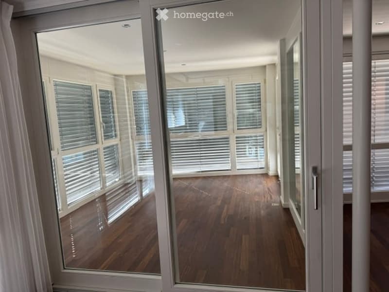 Empty room with wooden flooring and large glass sliding doors leading to an outdoor area. The room has white walls and large windows with blinds.
