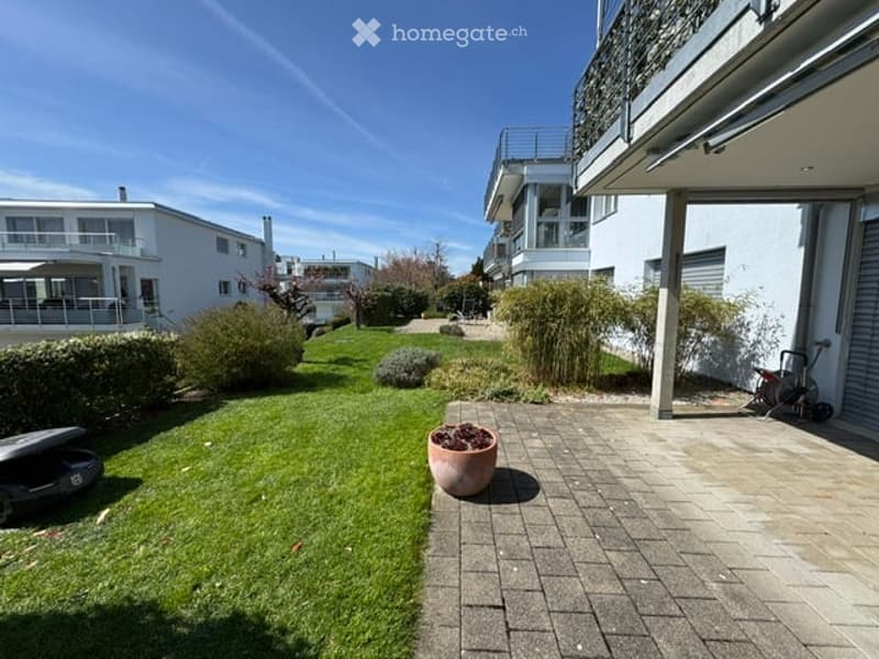 Well-maintained garden with a paved pathway leading to a large modern house. Features include neatly trimmed hedges, a large flower pot, and a stroller parked near the house.