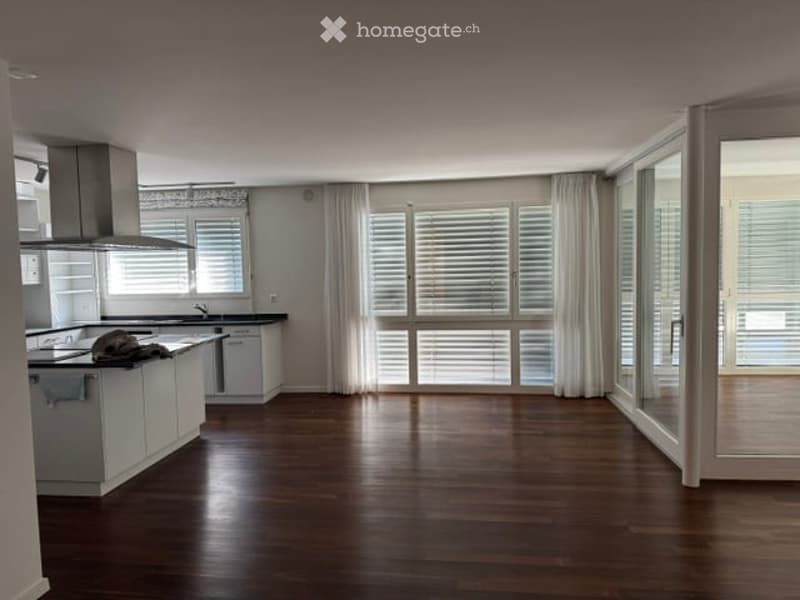 Modern kitchen with white cabinets, a central island, and large windows offering a view. The floor is made of dark wood, and the space is well-lit with natural light.