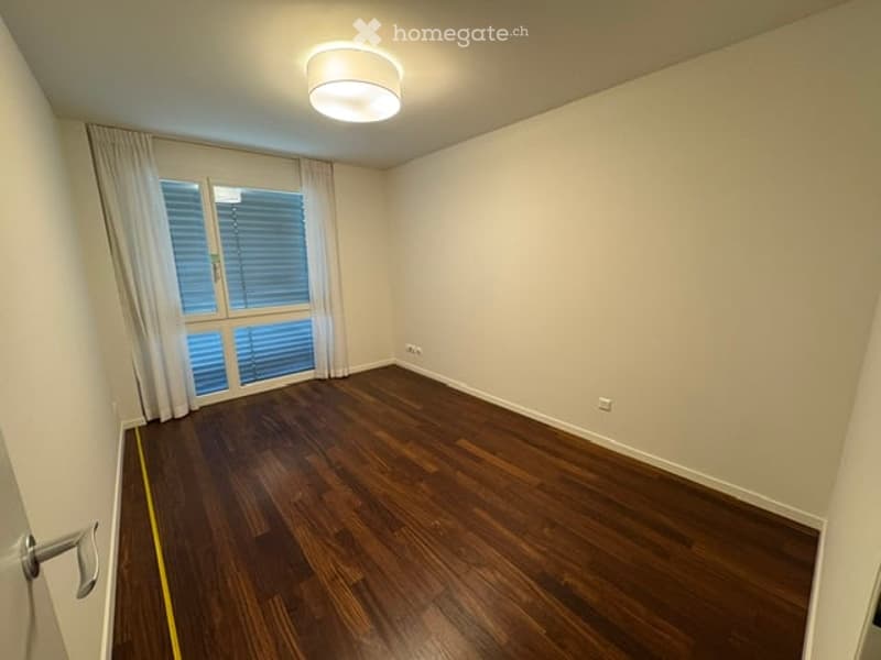 Empty room with wooden flooring, white walls, large window with blinds, and a ceiling light fixture.