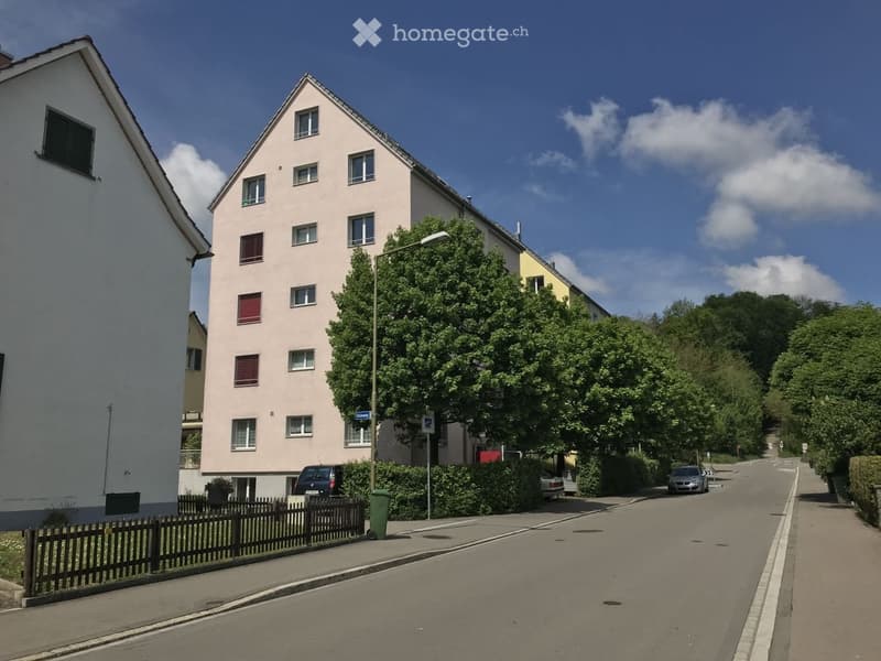 A multi-story residential building with light pink facade and multiple windows, situated on a quiet street with trees and parked cars.