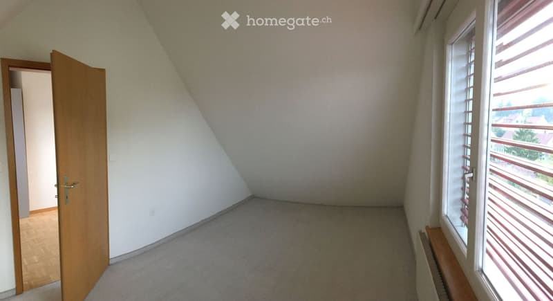 Empty attic room with wooden floor, carpeted area, large window with blinds, and open wooden door leading to another space.