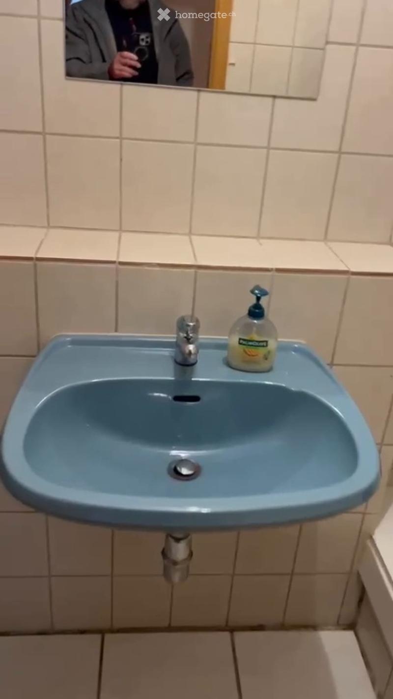 A bathroom with a blue sink, a silver faucet, and a soap dispenser on the counter. The walls are tiled in beige, and a mirror is mounted above the sink.