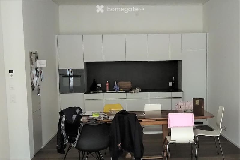 A modern kitchen with white cabinets, a wooden dining table, and several chairs. There is a black oven and various items on the counter.