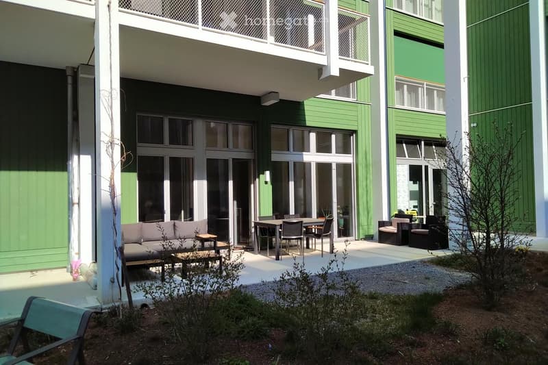 Outdoor seating area with a sofa, small table, dining table, chairs, and a bench, surrounded by greenery and a modern building with green panels and large windows.