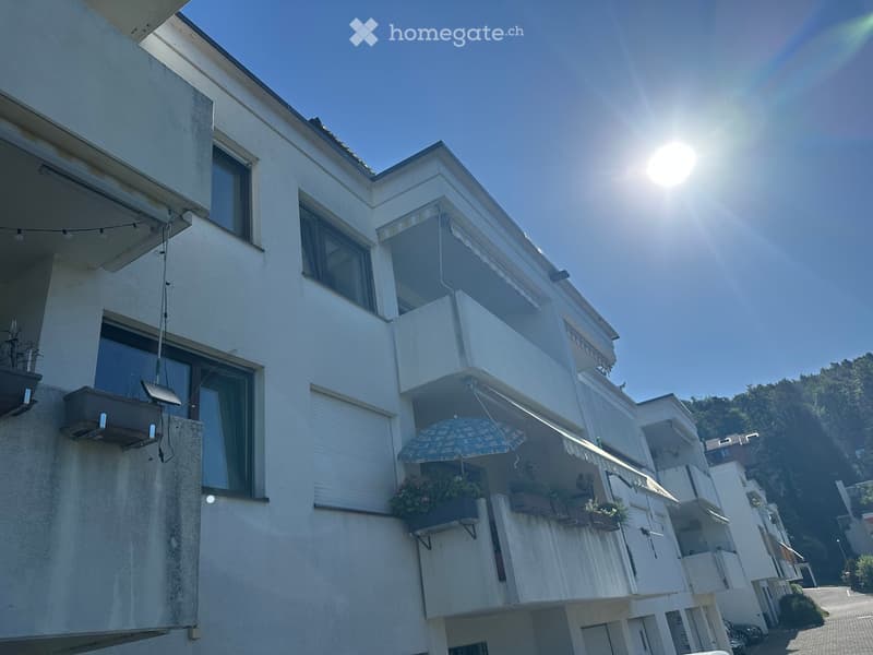Multi-story residential building with balconies, large windows, and outdoor furniture including an umbrella and potted plants.