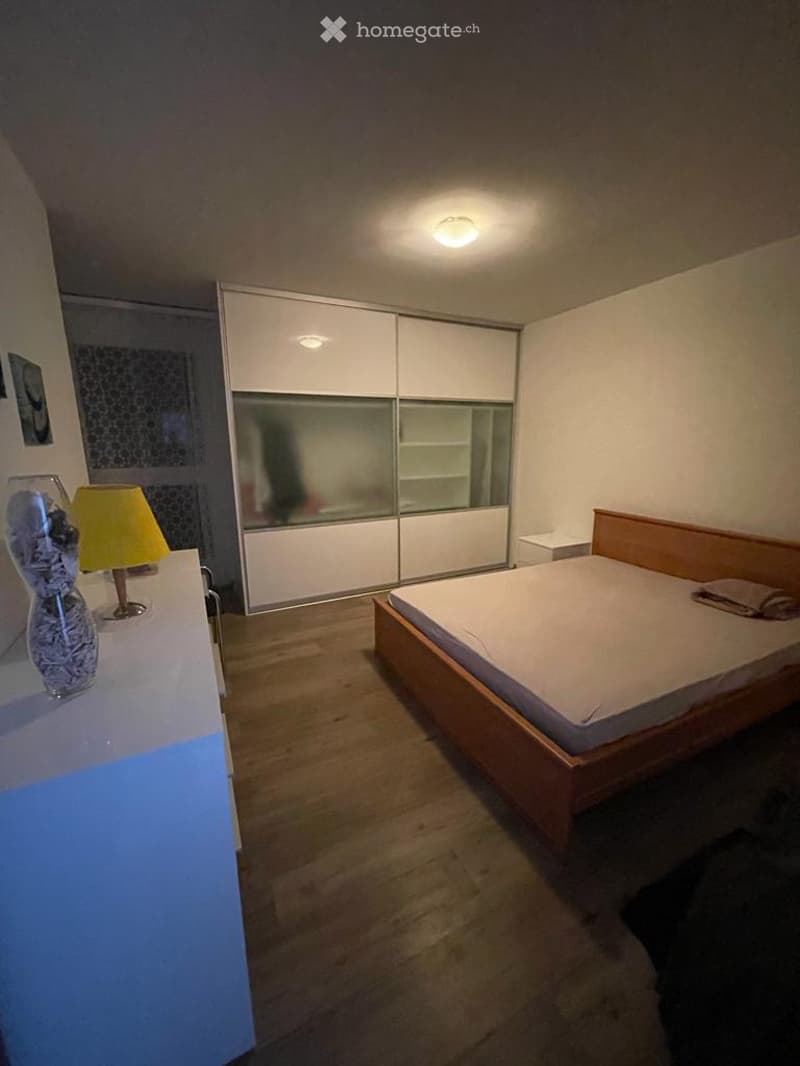 A modern bedroom with a wooden bed, white wardrobe, bedside lamp, and decorative vase on a nightstand.