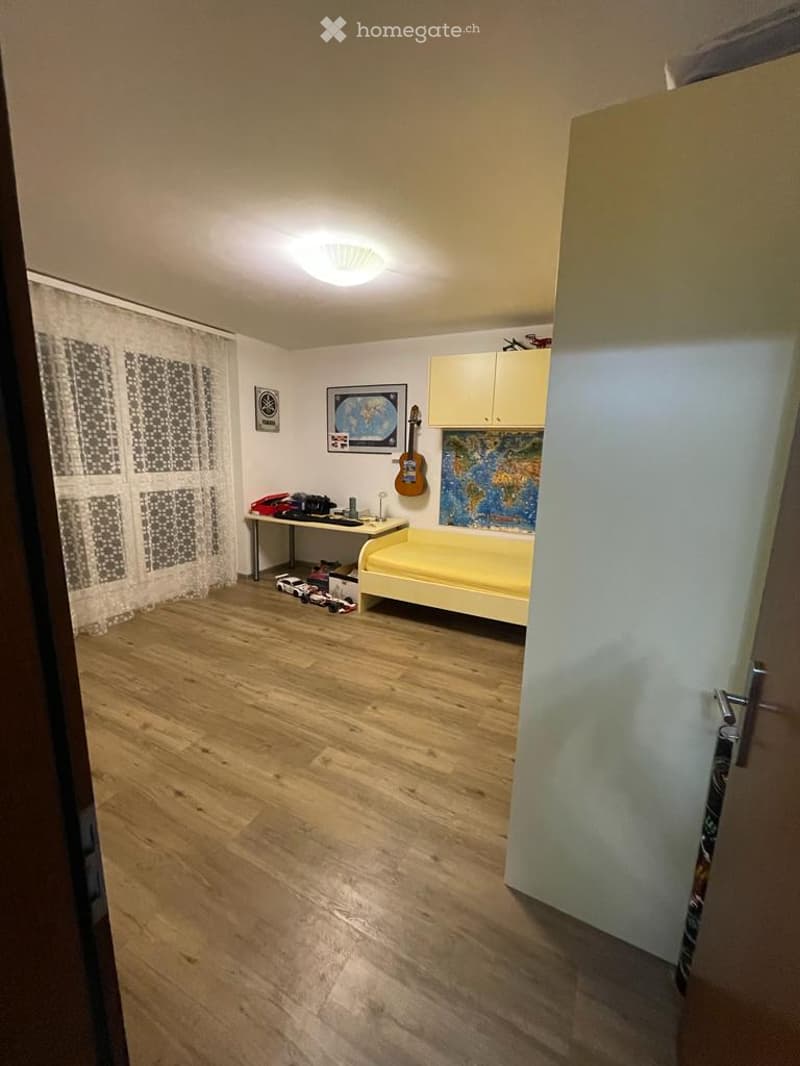 A small, well-lit bedroom with wooden flooring, a single bed, a small table with various items, a guitar on the wall, a cabinet, and a large window with a curtain.