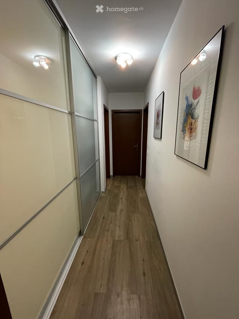 A modern hallway with wooden flooring, white walls, and recessed lighting. Features include a glass-paneled door on the left and framed artwork on the right.