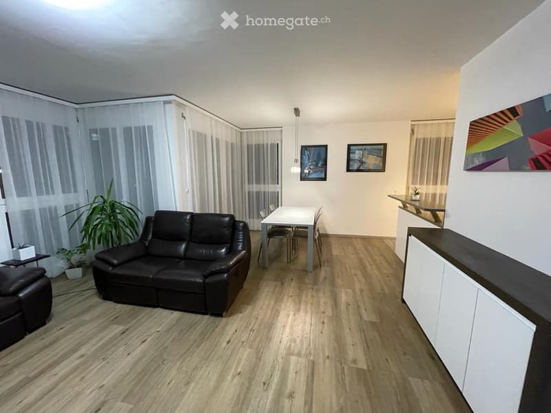 Modern living and dining area with a black leather sofa, a white dining table with chairs, a small cabinet, and a potted plant. The space features wooden flooring and large windows with white curtains.