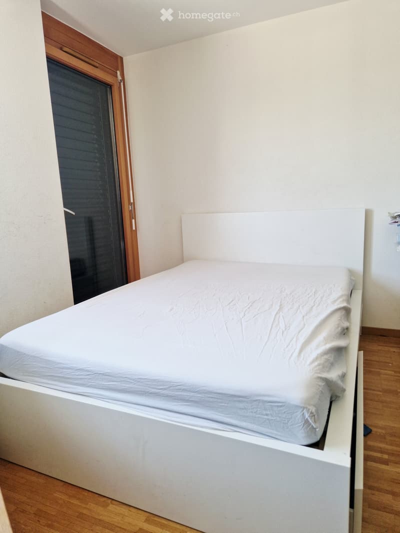 Minimalist bedroom with a white bed and headboard, wooden flooring, and a large window with blinds.
