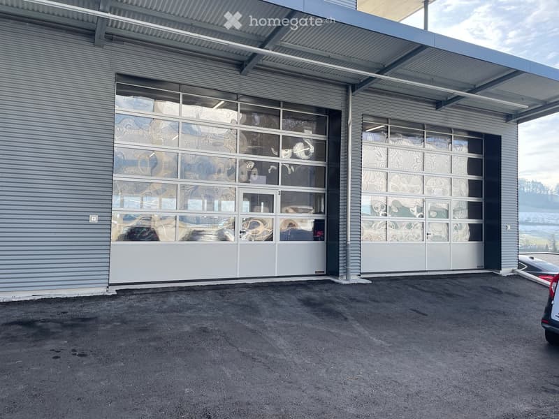 Modern industrial-style building with large garage doors and glass panels, featuring a covered entrance area and paved parking lot.