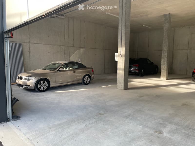 Underground garage with concrete walls and pillars, two parked cars, and a partially open garage door.
