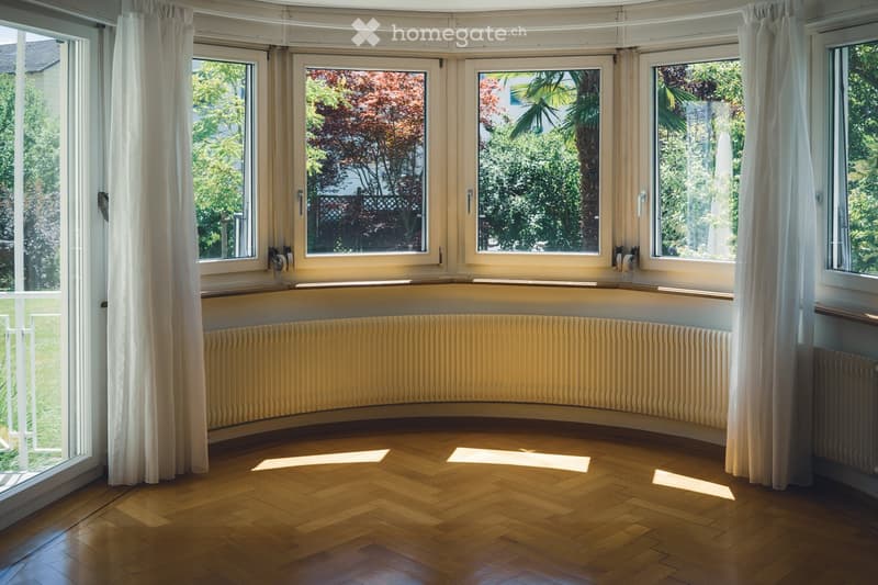 Empty room with wooden flooring, large windows with white curtains, and a radiator along the wall.