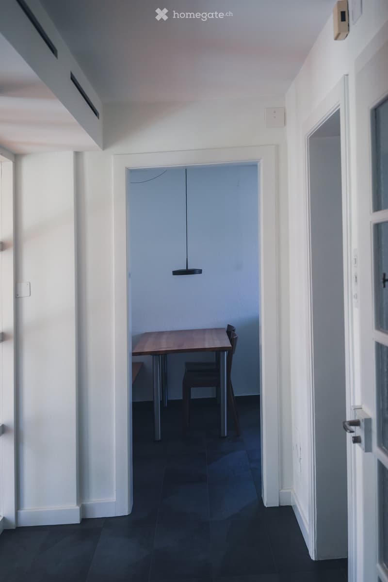 A modern, minimalist hallway with white walls, dark tiled flooring, and a pendant light hanging from the ceiling. An open doorway leads to a small room with a wooden table and chairs.