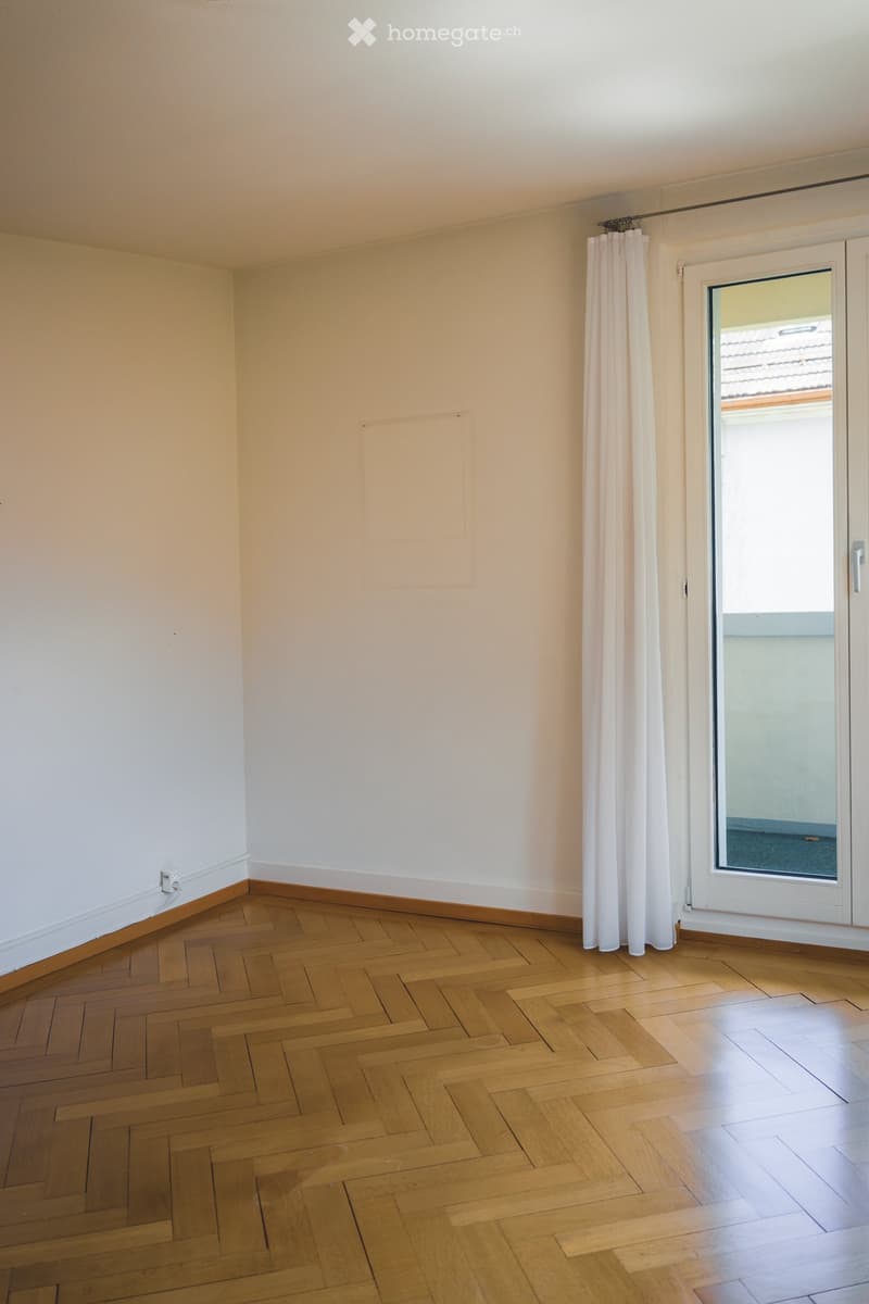 Empty room with wooden herringbone flooring, white walls, and a large window with white curtains.