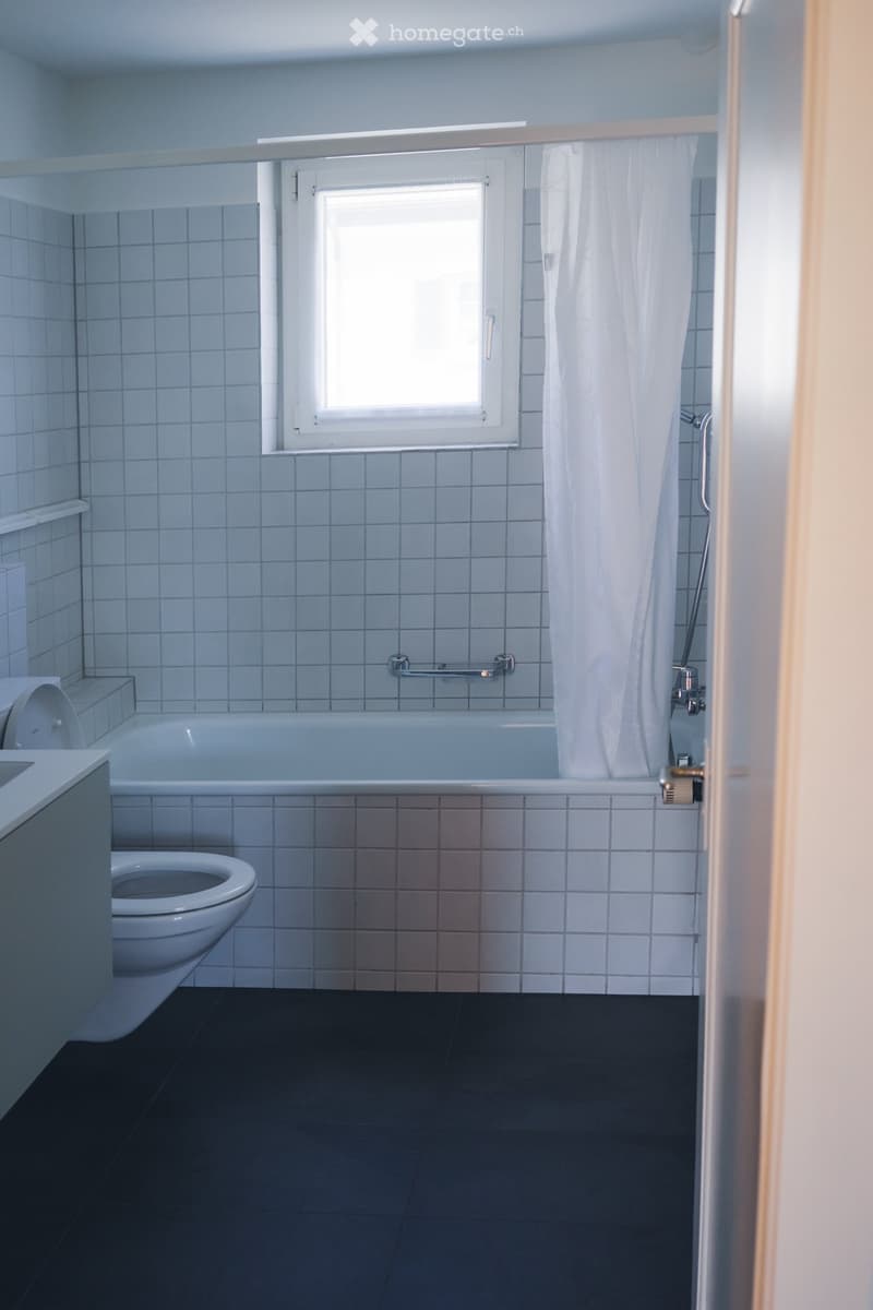 Modern bathroom with a bathtub, toilet, and shower curtain. The walls are tiled in light blue, and the floor is dark grey.