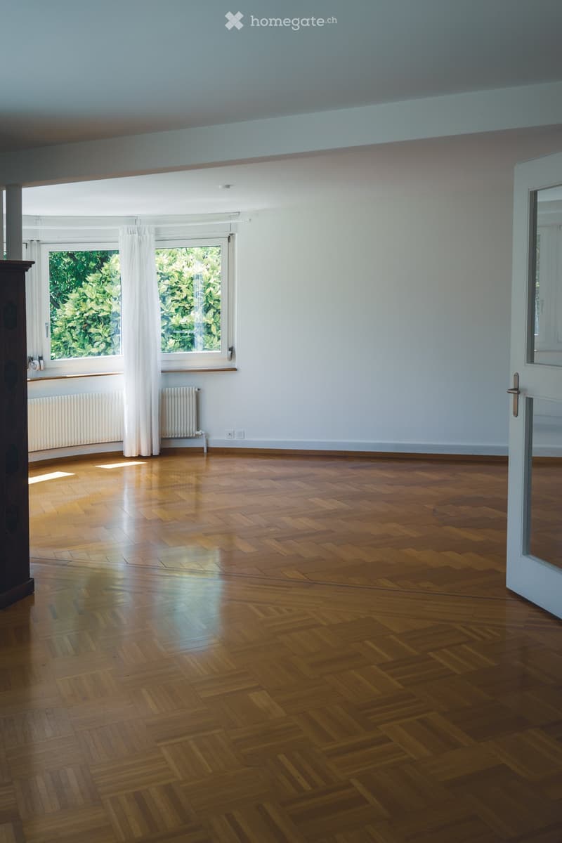 Empty room with wooden parquet flooring, white walls, large windows with white curtains, and radiators beneath the windows.