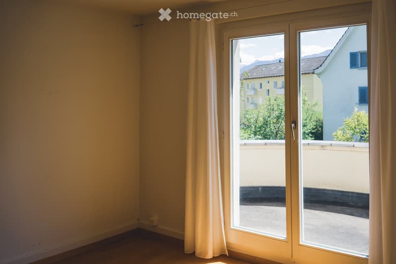 Empty room with wooden flooring, large window with white frame, and light-colored curtains.