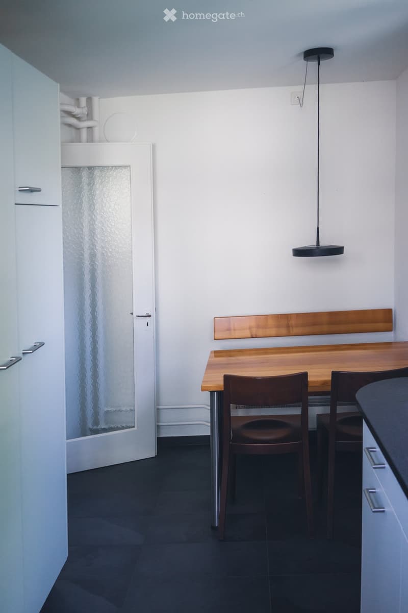 Modern, minimalist room with a wooden table, two chairs, a floor lamp, and a white cabinet with a frosted glass door.