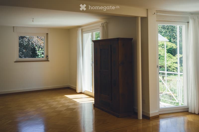 Empty room with wooden flooring, large windows with white curtains, and a tall wooden cabinet. Natural light streams in from the windows.