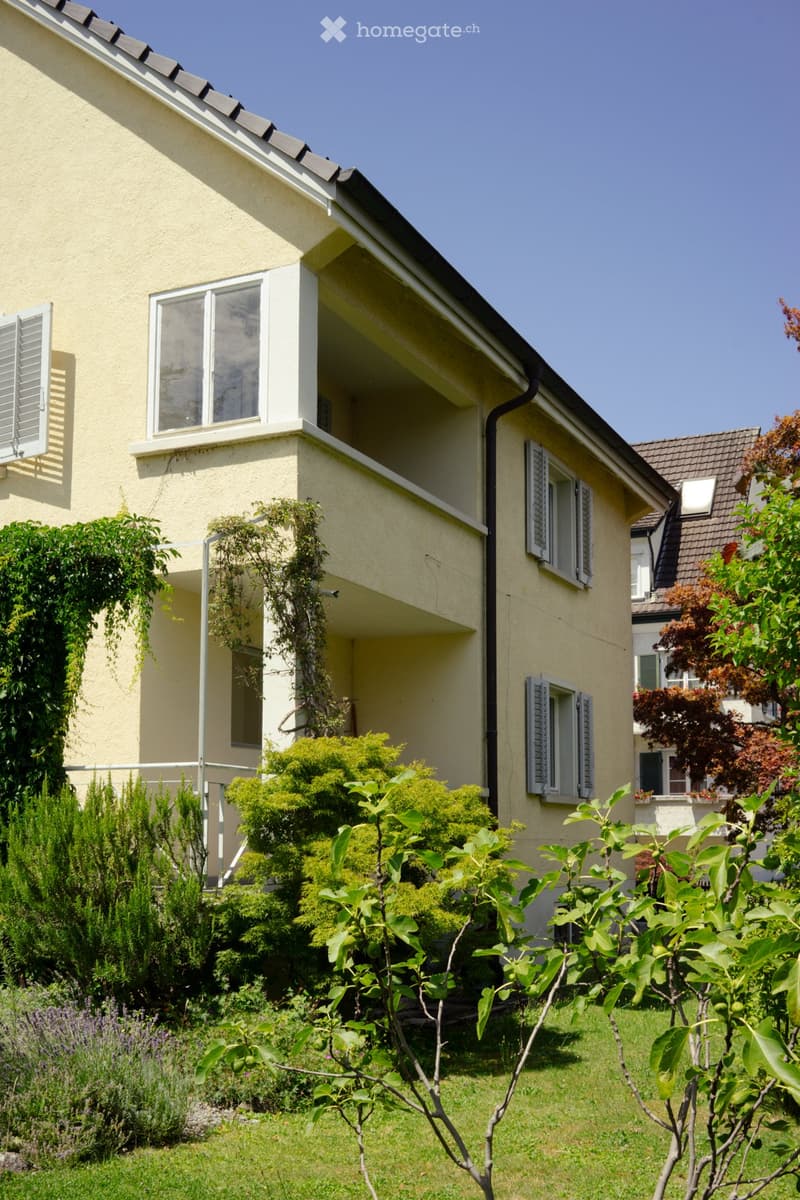 Two-story house with light yellow walls, multiple windows with shutters, and a well-maintained garden with various plants and trees.